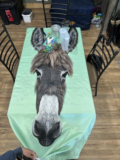 A dining room table with a mint green tablecloth emblazoned with a lifelike head of a donkey. The head is almost 6 feet tall: bigger than will fit on this table that easily seats six people.