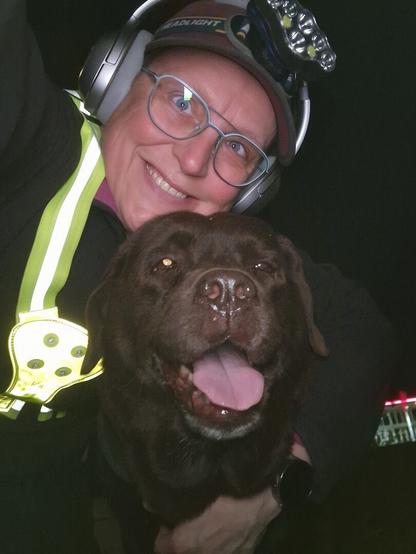 Night time selfie of a person and a chocolate brown Labrador with greying snout. The person is wearing a black jacket, yellow safety harness, red cap, silver headphones and blue-rimmed glasses. On their cap rests a headstrap torch in off position. Their free hand is around the dog's shoulders. Both are facing the camera. The dog's mouth is slightly open, showing off some blep. The person's head is resting on the dog's head. They're smiling, grateful for their time spent together.