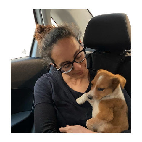Forografia a color de una humana con gafas negras sosteniendo a un cachorro de perro de color marrón naranja y pecas blancas en su cuello y en sus piernas. Están sentada en el asiento de atrás.