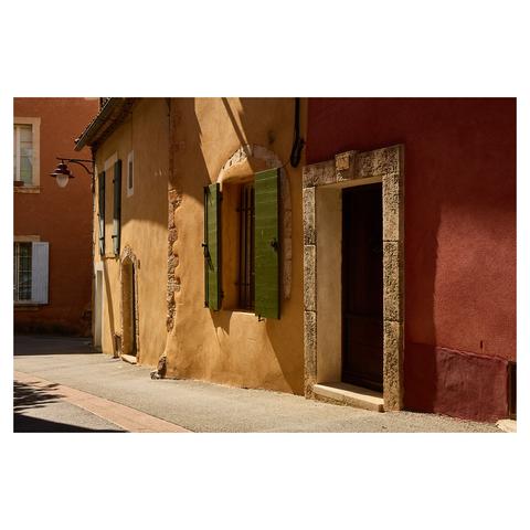 Tight, angled image of colourful facades in a village in Provence. The walls are painted in shades of ochre, with green wooden shutters. A streetlamp attached to the building wall is visible on the right. Warm sunlight creates long shadows.