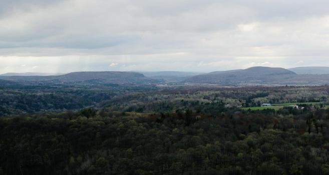 Photograph of a panoramic view of a hilly landscape under a cloudy sky, featuring varied shades of green, brown and red in the trees, along with open fields and distant hills. A green field with farm buildings can be seen in the distance. This could be an autumnal scene but it is spring, with surprisingly lots of colours in the landscape.