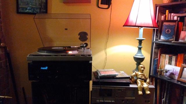 A photograph of a living room with gold painted walls. There is a stereo with a turntable and an album on it. The LED reads "PHONO". Next to the stereo is a lamp with a pink shade, and a miniature C3PO figurine, and to the right is a bookcase.