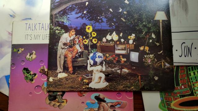 A collection of vinyl records stacked on top of each other. On top is World Party's "Private Revolution", the album cover depicting a man with brown curly hair and sunglasses playng a guitar. Around him are sunflowers, trees, a lamp, and a statue of Atlas holding up the planet Earth.