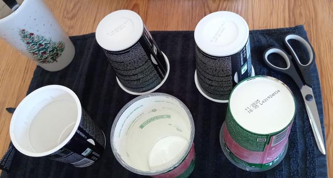 photo of five plastic cup-noodle pots with paper sleeves. they are empty and clean and have no lids. they are sitting on a towel on a wooden table with kitchen scissors next to them.