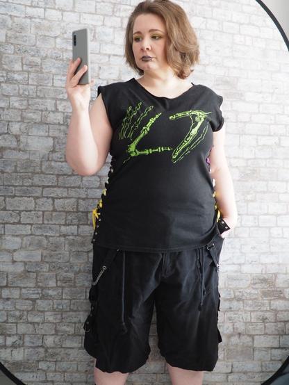 Photo of a chubby white woman shape wearing a black band t-shirt (bright green L7 stylised as skeleton hands print) with neckline and sleeves cut off. With long baggy shorts with chain straps.