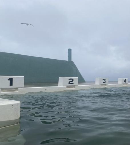 White concrete lap lane markers numbered 1 to 4 in black sit above dark green waters of the lap pool at the Baths. Behind them is a high turquoise concrete wall with a triangular end topped by a chimney like shape. A seagull is flying in the grey skies above the wall.