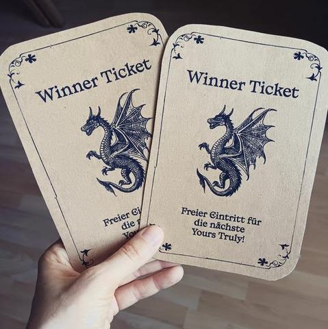 Ein Foto von meiner Hand, die zwei große, rechteckige Karten aus braunem Papier hochhält. Bedruckt sind sie in schwarz mit der Zeichnung eines Drachen. Über dem Drachen steht Winner Ticket und darunter: Freier Eintritt für die nächste yours truly.