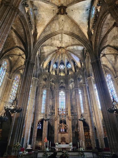 The tall stone columns of the church choir with the altar at center and stained glass windows glowing brightly. the pillars support a ceiling with ribbed vaulting.