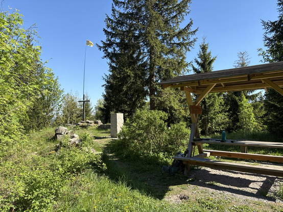 A portable antenna mast attached to a summit cross with a wire antenna leading to a bench, large trees in the background.