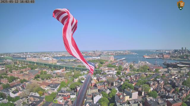 Flag in the wind, from the top of Bunker Hill Monument. Green trees and buildings below, with the waters of Boston Harbor in the distance, all under sunshine and blue skies today. This image is from one of the four National Park Service web cameras located at this Boston National Historical Park site.