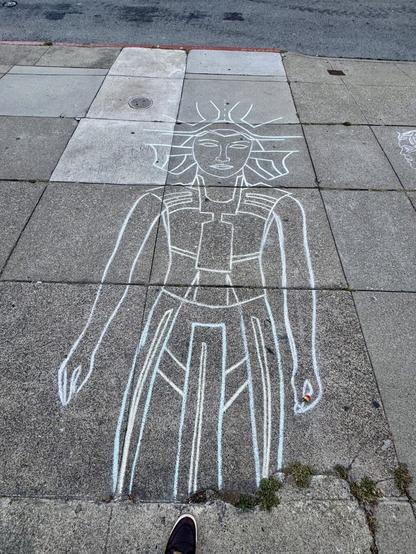 A chalk drawing on the sidewalk. It's about 6 feet tall and 4 feet wide. It's a drawing of a person in a style that reminds me of ancient Egyptian art, except the head is turned toward the viewer. The art is very stylized and drawn in white and blue chalk.