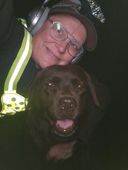 Night time selfie of a person and a chocolate brown Labrador with greying snout. The person is wearing a black jacket, yellow safety harness, red cap, silver headphones and blue-rimmed glasses. On their cap rests a headstrap torch in off position. Their free hand is around the dog's shoulders. Both are facing the camera. The dog's mouth is slightly open, showing off some blep. The person's head is resting on the dog's head. They're smiling, grateful for their time spent together.