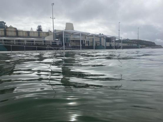 Taken while swimming in the baths, looking across the water to the building and shade structures on a cloudy grey day. Just enough light reflecting on the water surface to make it shine like silk.