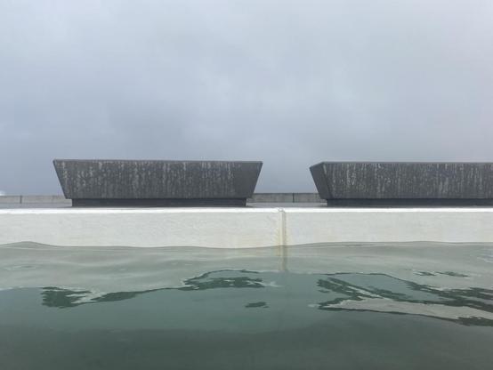 Above green water and the white edge of the main pool, two solid grey concrete benches sit on the concourse streaked darker with rain.