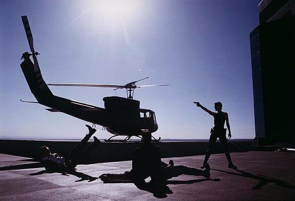 Still from the movie "The Matrix" of Trinity (Carrie Anne Moss) standing on the roof of a building and silhouetted against the sky, holding a pistol, as a UH-1 Huey helicopter approaches.