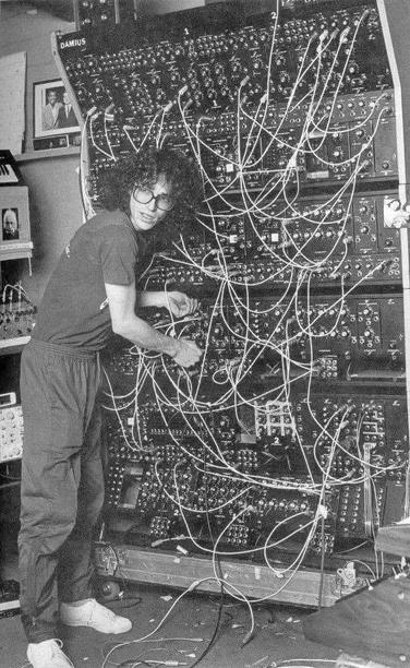 The image depicts a person standing next to a large, complex electronic device composed of numerous interconnected components and wires. The individual, wearing glasses, a t-shirt, and sweatpants, appears to be interacting with the machine. The setting includes framed pictures and other electronic equipment, suggesting a workspace or laboratory environment. The intricate wiring and components indicate that this could be an early computer or a sophisticated piece of electronic equipment, highlighting the complexity of technology in its early stages.