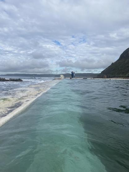 Looking southwards along the back wall of the baths. The white of the wall and nearby floor makes the a lovely wide pale turquoise line in the water before giving way to a deeper green. Sky is full of white clouds with only a few small patches of blue.