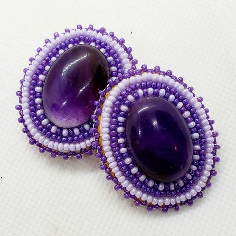 a pair of oval-shaped beaded earrings by kiamichi. the centerpiece is a large translucent deep purple gemstone surrounded by shiny white and purple beads
