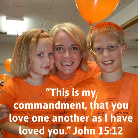 A smiling woman poses with a girl and a boy, both wearing orange shirts. The background features orange balloons. Text overlaid on the image quotes John 15:12 about love.