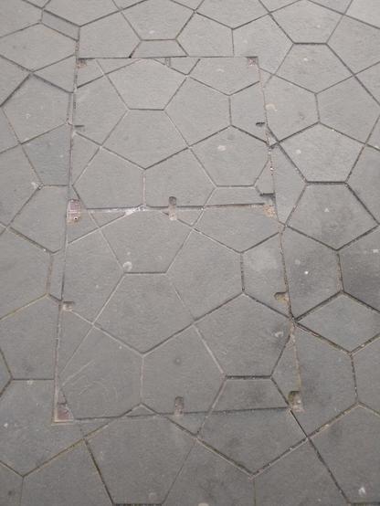 A photograph of street paving made of pentagons. In the middle are two square areas: the bottom square has tiles that mostly match the surrounding tiling, but the upper square has been made to fit leaving awkward tiny tiles that don’t match.