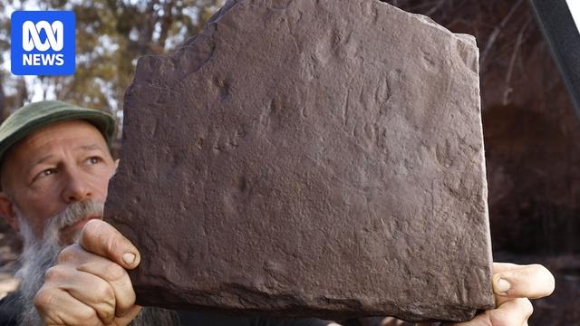 Man with grey beard and hat (Craig Eury) holds up rock with fossil tracks in it.