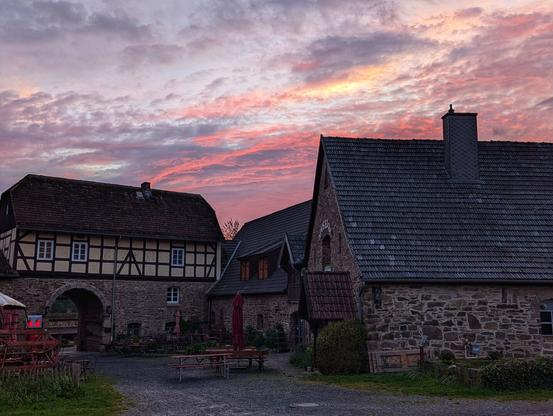 A ground-level, medium shot captures a collection of old, stone and timber-framed buildings beneath a vibrant sunset sky. The sky is a blend of pinks, purples, and oranges, with textured clouds. The buildings are constructed from dark stone on the lower levels and timber framing with white infill on the upper levels, all topped with dark tiled roofs. An arched gateway is visible in the leftmost building. A gravelled courtyard lies in front of the buildings, featuring a wooden picnic table and some greenery. The overall lighting suggests dusk.