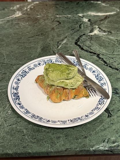 Croissant topped with green cream on a floral plate with cutlery.