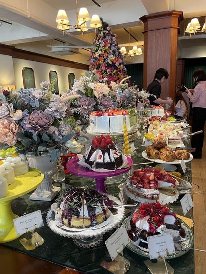 A dessert buffet table displays a variety of cakes, including strawberry, blueberry, and cherry chocolate options. Floral arrangements add a decorative touch.