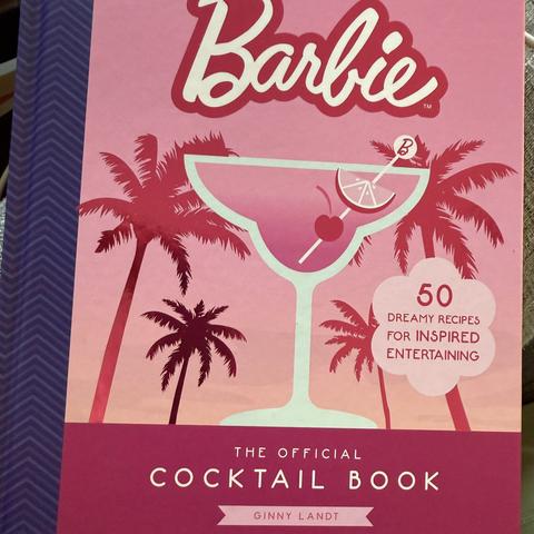 Pink hardcover book with purple binding. At the top is the Barbie logo. Below that is pink cocktail in a glass, against a background of pink palm trees against a (you guessed it) pink sunset. In the lower right it says “50 dreamy recipes for inspired entertaining.” At the bottom it says The Official Cocktail Book, Ginny Landt.