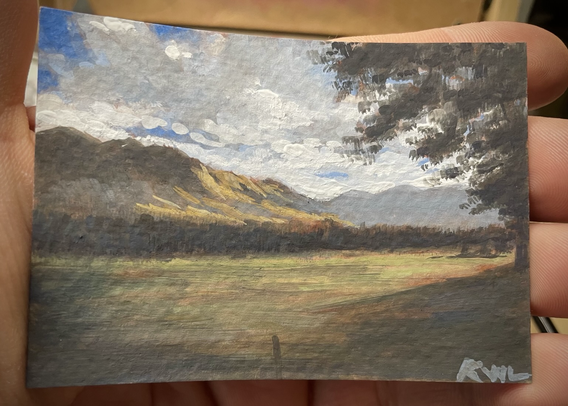 A miniature painting, held in the palm of a hand, of a large mountain dramatically lit with trees and a great field in front of it and bright clouds in the sky.