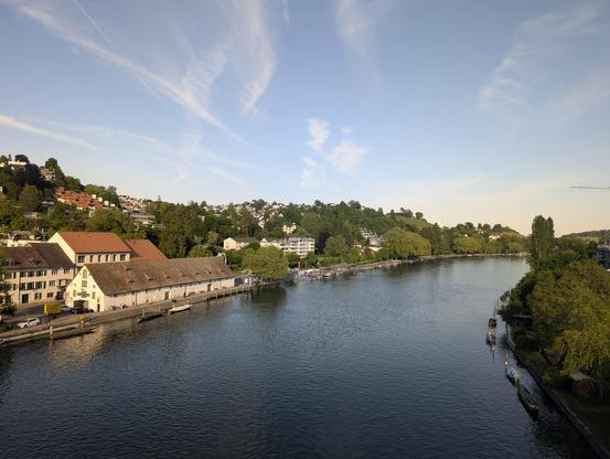 Blick ab einer Brücke auf einen abendlichen Fluss. Links ein langes Gebäude mit Ziegeldach (und weiter Gebäude, dahinter grüne Hügelkette), rechts bewaldetes Ufer