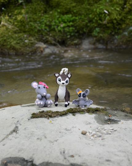 Trois amigurumis crochetés sont posés sur un rocher au bord d'une rivière. On voit une souris grise avec un nœud rose, un cerf marron et blanc et une chauve-souris grise avec un nœud orange. Derrière eux, de la mousse verte couvre les rochers et l'eau coule doucement. L'ambiance est calme et naturelle.