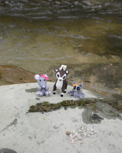 Trois amigurumis crochetés sont posés sur un rocher au bord d'une rivière. On voit une souris grise avec un nœud rose, un cerf marron et blanc et une chauve-souris grise avec un nœud orange. Derrière eux, de l'eau coule doucement. L'ambiance est calme et naturelle. La vue est prise légèrement au-dessus d'eux