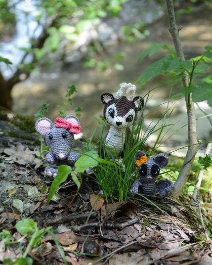 Trois amigurumis crochetés sont posés dans la nature. On voit une souris grise avec un nœud rose, un cerf marron et blanc et une chauve-souris grise avec un nœud orange. Ils sont entourés de végétation.  Derrière eux, on devine la rivière qui coule en contrebas. L'ambiance est calme et naturelle.