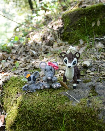 Trois amigurumis crochetés sont posés sur un roche plein de mousse, dans la nature. On voit une souris grise avec un nœud rose, un cerf marron et blanc et une chauve-souris grise avec un nœud orange. Derrière eux, il reste des traces de cet automne avec des feuilles mortes gisant sur le sol. L'ambiance est calme et naturelle.