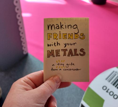 A small folded-up zine in the hand of a white woman with long, unpainted finger nails. In the background you can make out a hot pink carpet, grey flooring, and something that rightly looks like a giant archival box (because it is). The zine reads "Making friends with your metals: a tiny guide from a conservator" in various styles of handwriting.