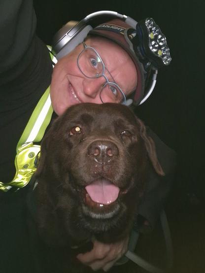 Night time selfie of a person and a chocolate brown Labrador with greying snout. The person is wearing a black jacket, yellow safety harness, red cap, silver headphones and blue-rimmed glasses. On their cap rests a headstrap torch in off position. Their free hand is around the dog's shoulders. Both are facing the camera. The dog's mouth is slightly open, showing off some blep. The person's head is resting on the dog's head. They're smiling, grateful for their time spent together.