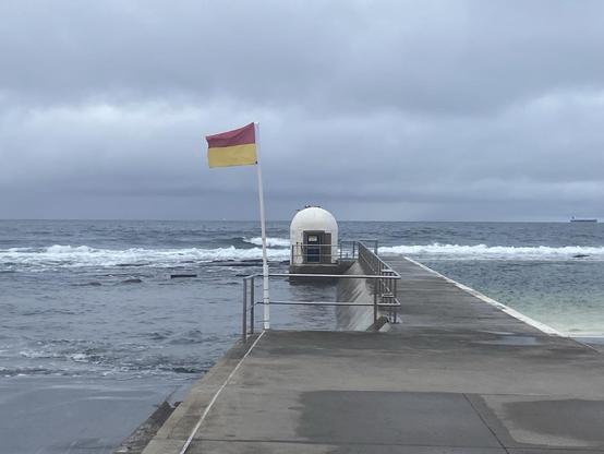 Looking along the northern concourse of the baths to the rounded white pump house and the ocean. The red and yellow lifesaving flag is up and fully stretched out by the southerly wind. Grey clouds fill the sky to the ocean horizon. The water looks greyer to match.