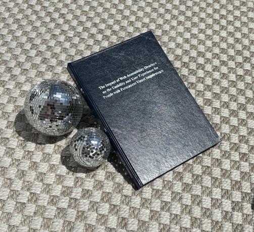 The printed version of the thesis titled "The impact of Web Accessibility Overlays on the Usability and User Experience for People with Permanent Visual Impairments", laying on a carpet next to two tiny disco balls.
