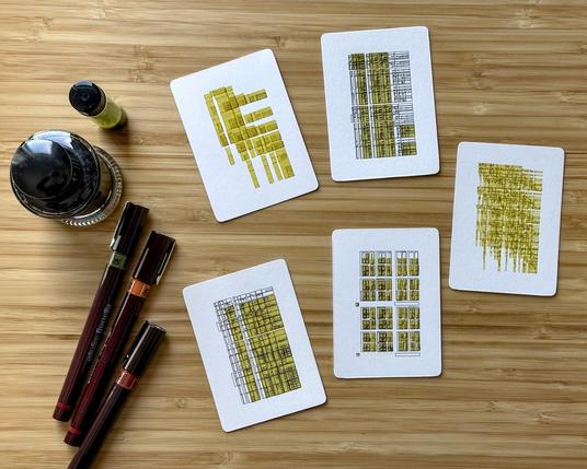 A bamboo desk with five small works of art on them, each 63mm x 88mm. They are in black and yellow, and the artwork is abstract line work based on a recursive algorithm that subdivides space in a regular way. The results are reminiscent of technical drawings. There are three Rotring Isograph pens on the left side of the trading cards, and two bottles of ink above them.
