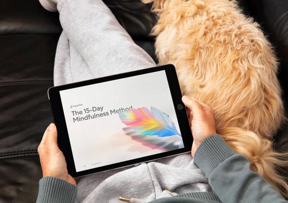 A person relaxes on the couch holding a tablet displaying "The 15-Day Mindfulness Method," with a fluffy dog nearby.
