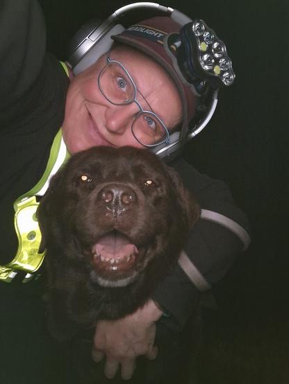 Night time selfie of a person and a chocolate brown Labrador with greying snout. The person is wearing a black jacket, yellow safety harness, red cap, silver headphones and blue-rimmed glasses. On their cap rests a headstrap torch in off position. Their free hand is around the dog's shoulders. Both are facing the camera. The dog's mouth is slightly open, showing off some blep. The person's head is resting on the dog's head. They're smiling, grateful for their time spent together.