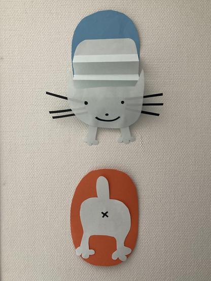 A bit of cat wall-art: a vertically-installed, two-piece, craft paper cat posted on a white wall. The top part is a greyish-white smiling cat with black whiskers and two small paws (coming out of the bottom of its head) emerging accordian-bodied out of an blue oval "portal". The bottom part is the rear end of the greyish-white cat disappearing into an orange oval "portal", short and stubby tail erect, two short legs, and a black "x" to designate the asshole. Artist: teenage daughter.