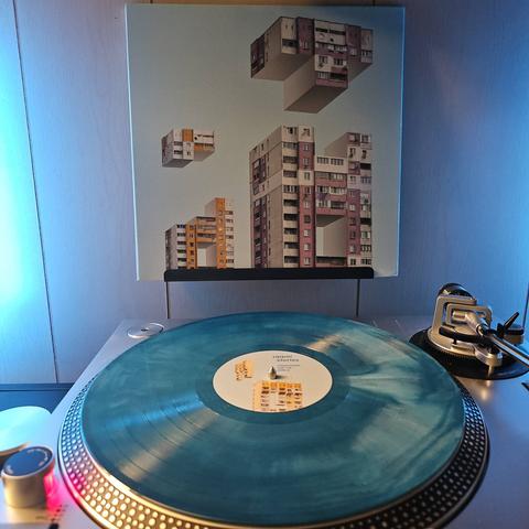 A Seafoam (Aquamarine and White Merge) vinyl record sits on a turntable. Behind the turntable, a vinyl album outer sleeve is displayed. The front cover shows apartment buildings missing pieces, and Tetris style pieces of buildings coming down from the sky.