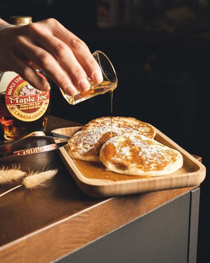 A hand pours maple syrup onto pancakes dusted with powdered sugar, presented on a wooden plate.