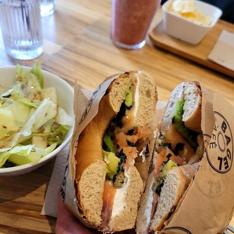 A bagel sandwich cut in half with cream cheese, salmon, avocado, and capers, alongside a salad, smoothie, and butter.