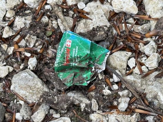 Eine Verpackung von etwas Undefinierbarem, vermutlich etwas mit Schokolade, liegt auf einem Waldweg.