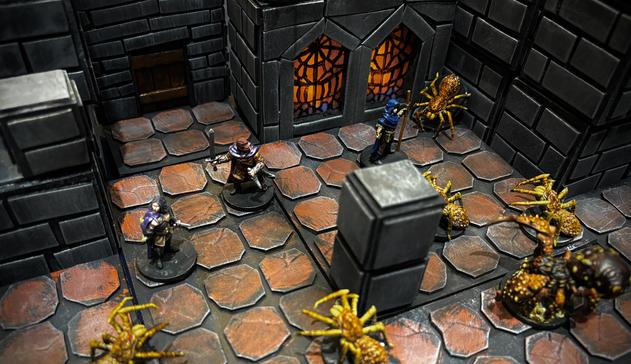 A photo of a painted miniature adventuring party encountering some spiders in a dungeon. A fighter holding a sword, a rogue holding a dagger, and a Druid holding a staff are bracing themselves for the attack from multiple monstrous spiders. In the background is the dungeon scenery including a doorway and a pair of arches with stained glass windows.