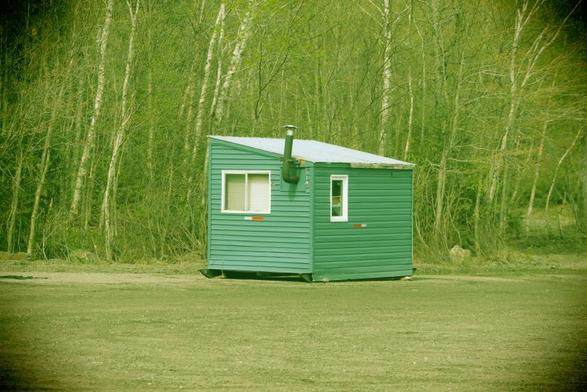 A green panelled ice hut in good condition, with a slanted roof, aluminum window and door, sits in the Spine Beach parking lot. Behind the ice hut is a mix of trees starting to show early Spring budding. The yellowy tinged green tone and vignetting emulates a plastic camera (in camera effect).
