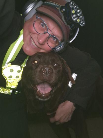 Night time selfie of a person and a chocolate brown Labrador with greying snout. The person is wearing a black jacket, yellow safety harness, red cap, silver headphones and blue-rimmed glasses. On their cap rests a headstrap torch in off position. Their free hand is around the dog's shoulders. Both are facing the camera. The dog's mouth is slightly open, showing off some mlem. The person's head is resting on the dog's head. They're smiling, grateful for their time spent together.